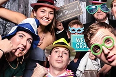 The Bride and Groom posing for photos on the Mirror Me, with their guests, in front of a backdrop, holding signboards and props