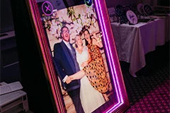 The Bride and Groom posing for photos on the Mirror Me, with their guests, in front of a backdrop, holding signboards and props