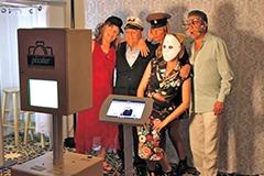 The Bride and Groom posing for photos on the Mirror Me, with their guests, in front of a backdrop, holding signboards and props