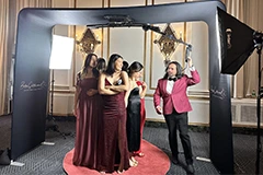 The Bride and Groom posing for photos on the Mirror Me, with their guests, in front of a backdrop, holding signboards and props