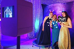 The Bride and Groom posing for photos on the Mirror Me, with their guests, in front of a backdrop, holding signboards and props