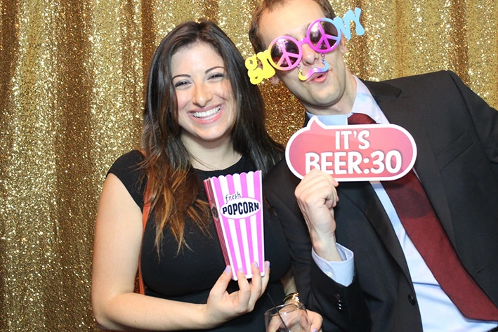The Bride and Groom posing for photos on the Mirror Me, with their guests, in front of a backdrop, holding signboards and props