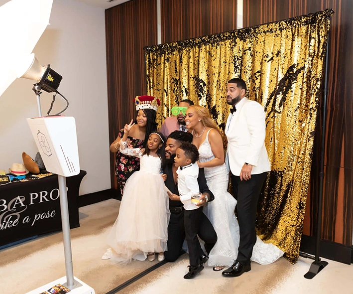 The Bride and Groom posing for photos on the Mirror Me, with their guests, in front of a backdrop, holding signboards and props