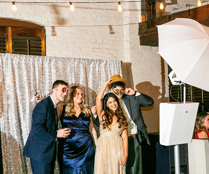 The Bride and Groom posing for photos on the Mirror Me, with their guests, in front of a backdrop, holding signboards and props