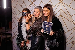 The Bride and Groom posing for photos on the Mirror Me, with their guests, in front of a backdrop, holding signboards and props