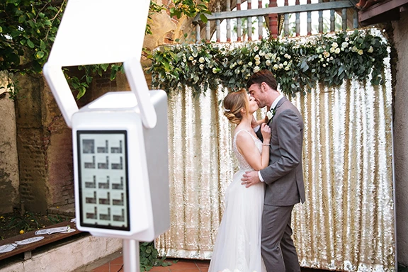 Couple kissing by a gold sequin backdrop with greenery at an anniversary photo booth rental.
