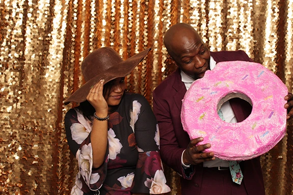 Holiday party photo booth: Woman in hat, man with donut prop against gold sequin backdrop.