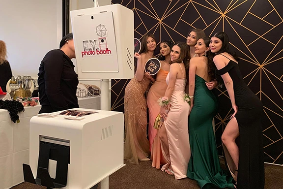 Group of young women in formal dresses pose at a photo booth with "EYE Photo Booth" visible.