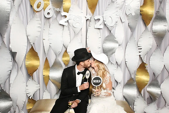 Couple kissing in front of gold and silver backdrop with wedding date