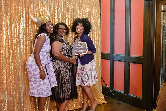 Three women posing at a photo booth for corporate events, with a gold backdrop and fun props.