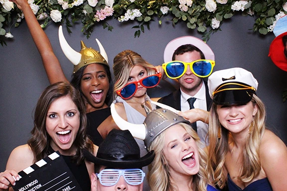 Group of friends posing with fun props at a selfie photo booth, smiling and laughing.