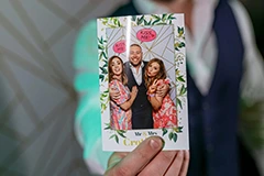 The Bride and Groom posing for photos on the Mirror Me, with their guests, in front of a backdrop, holding signboards and props
